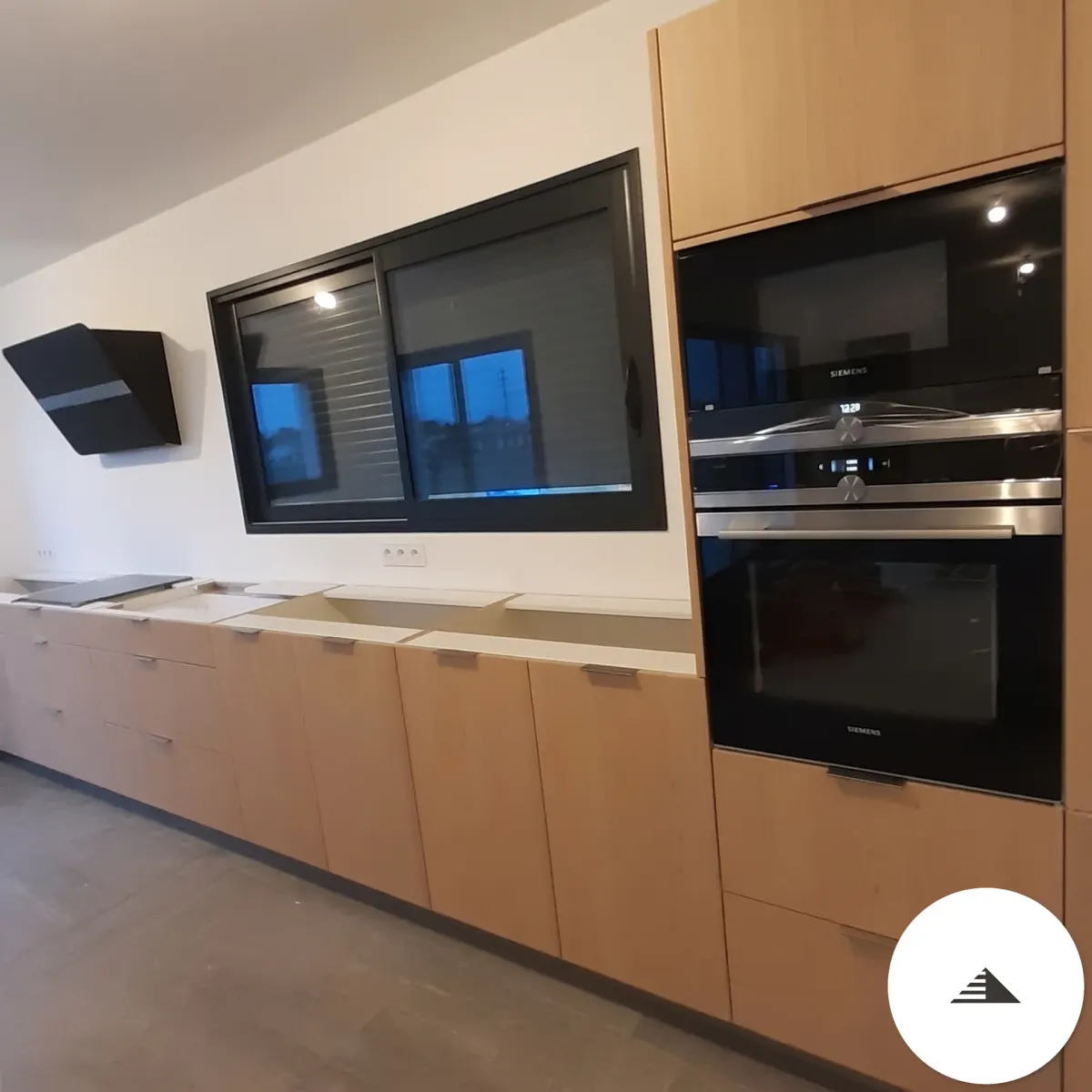 Cuisines sur mesure — Vue d'ensemble d'une cuisine en bois naturel clair avec colonne haute intégrant four et micro-ondes Siemens, hotte noire et sol béton gris à Toulouse | Gonzalez Agencement